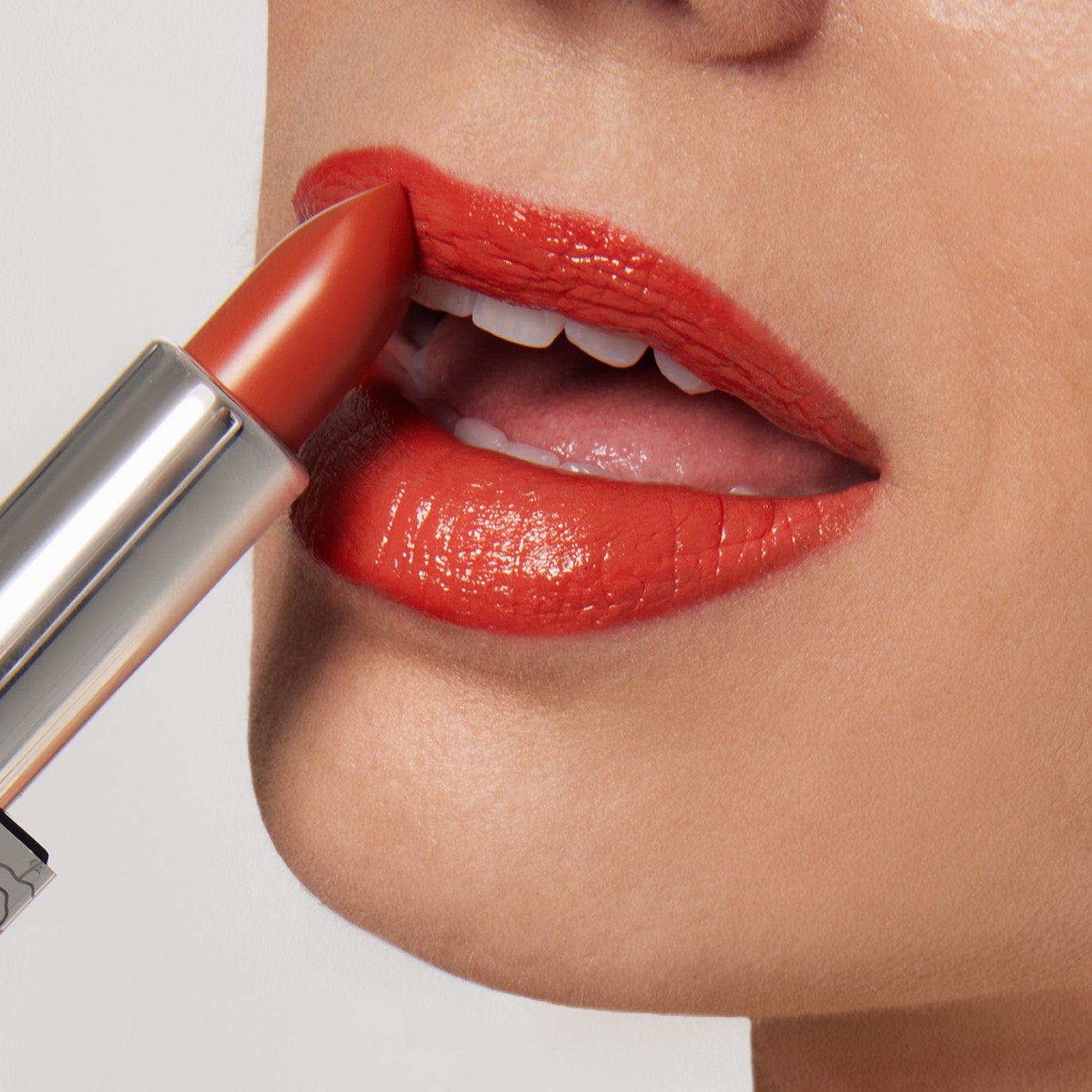A close-up captures a person applying Shopgalaxyss Beach House Shiny Velvet Cream Lipstick #122, in a bright orange hue with hydrating shea butter. Their parted lips reveal teeth, showcasing the vegan lipstick’s shiny finish against a soft, light background.