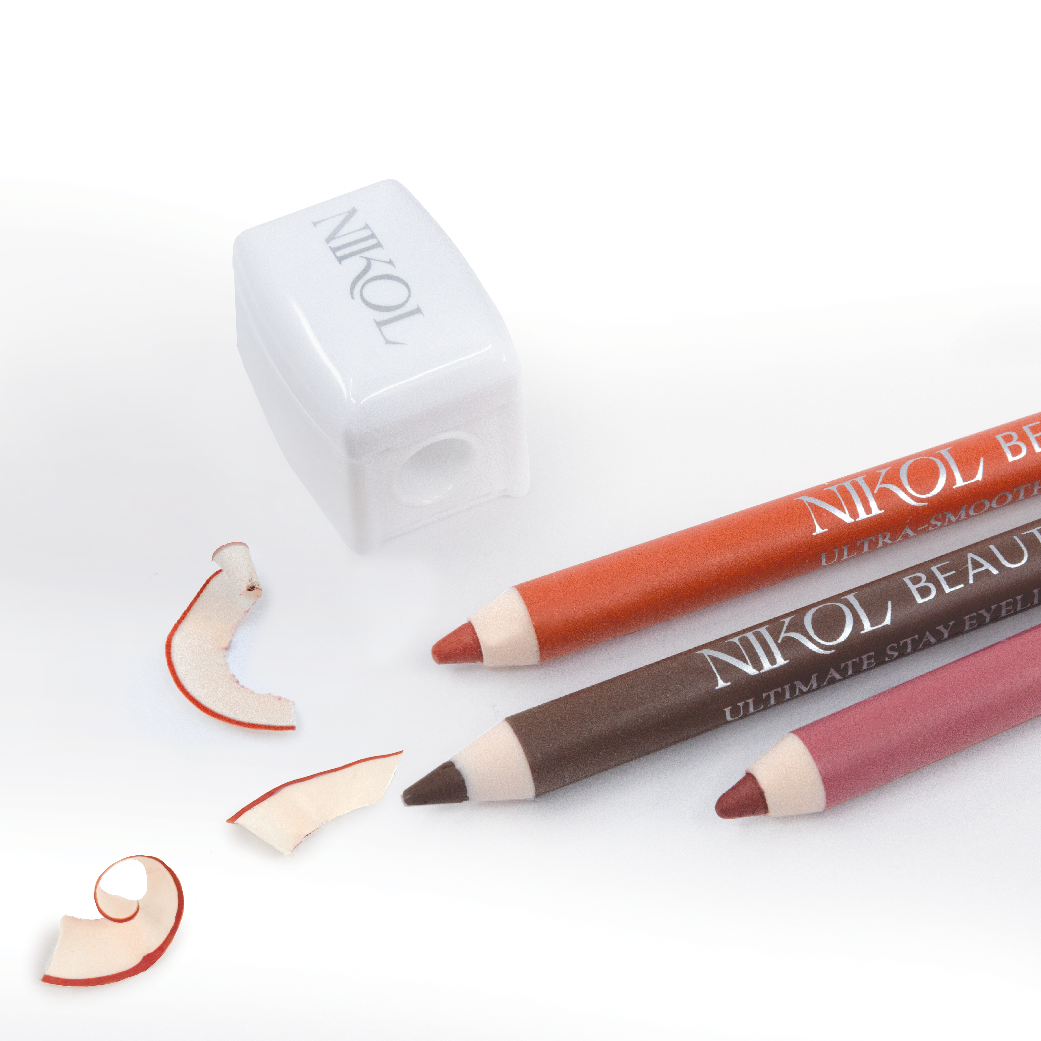 Two Shopgalaxys eyeliners, in brown and red, are elegantly placed beside a Pencil Sharpener on a white surface. Scattered around are shavings that have red-tinged edges, showcasing professional application—a true makeup bag essential.