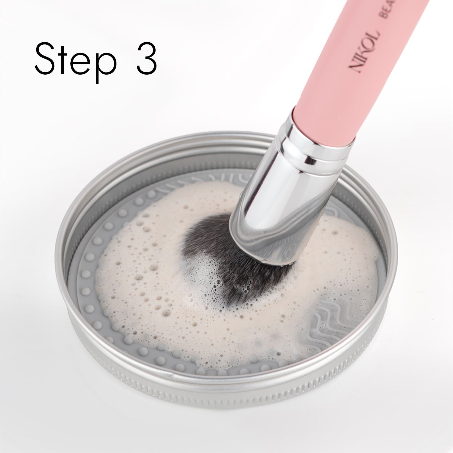 A makeup brush with a pink handle and silver ferrule is cleaned in a lid filled with Shopgalaxyss Lux Brush Soap infused with soapy water. Bubbles dance on the surface, while Step 3 in the top left corner ensures your beauty routine stays pristine.