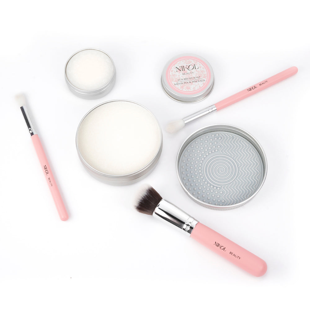 Nikol Beautys makeup brush cleaning kit includes pink-handled brushes, a textured pad, and tins of Lux Brush Soap with coconut oil, all artfully set against a white backdrop.
