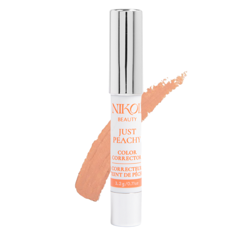 A tube of Shopgalaxyss Just Peachy Color Corrector, created for undereye discoloration, stands against a white background, with a peach-colored swipe behind it. The tube shows metallic accents and the products name and weight.
