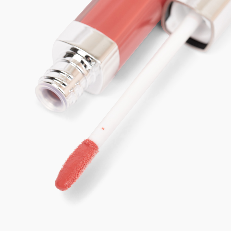 Nikol Beautys New Beauty Lip Gloss #282 offers high-impact coral pink shine. Featuring a white wand and soft applicator, this gloss includes nourishing avocado oil and is capped with shiny silver. It sits elegantly against a plain white background.