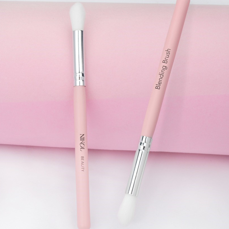 On a pink and white gradient background are two pink makeup brushes with white bristles. One handle reads Shopgalaxys, while the other, labeled Blending Brush, is ideal for mature eyes. Featuring silver ferrules, both are crafted from 100% vegan materials by Shopgalaxys.