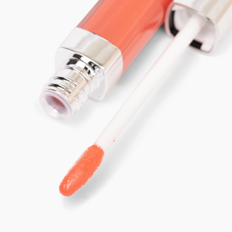 A close-up of the Defiance Lip Gloss #62 applicator from Shopgalaxys dipped in coral pink non-sticky gloss. The angled applicator is directed towards the viewer, with the tube seen in the background. This clean beauty product offers a shiny, smooth texture for high-impact shine.