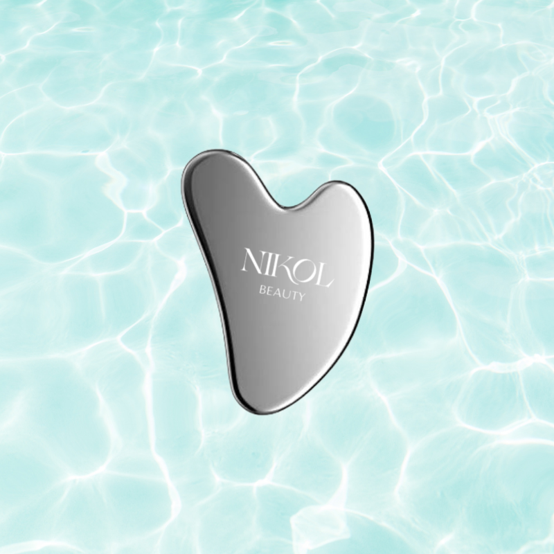 A Gua Sha Stainless Steel tool from Shopgalaxys, engraved with the brands name, rests against a backdrop of calm, light blue rippling water, designed to boost lymphatic drainage.